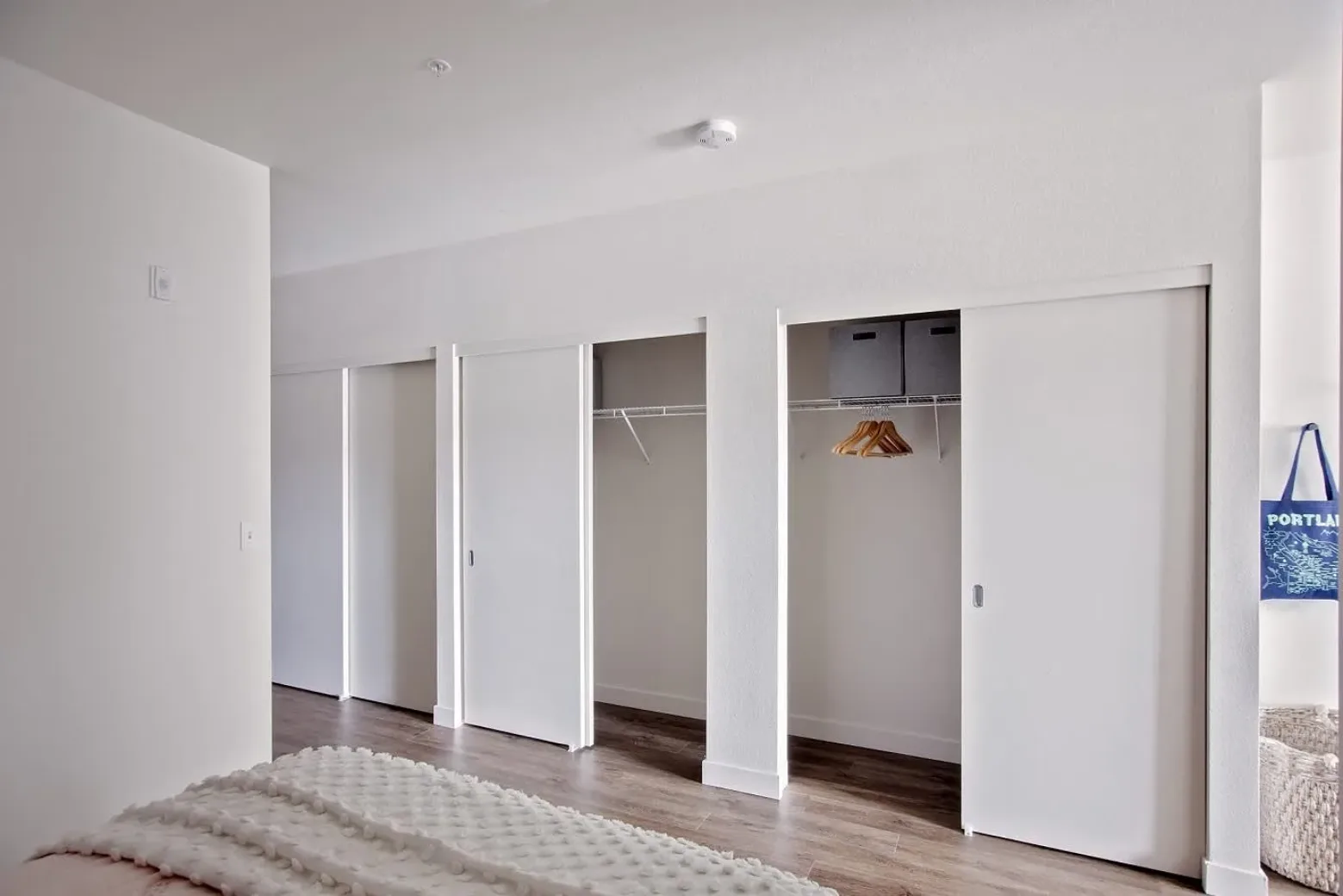 Interior of an apartment showing a bedroom with multiple sliding closet doors, open to reveal empty shelves, hangers, and storage boxes. A bed with a textured white blanket is visible in the foreground, set against a wall with light wood flooring.