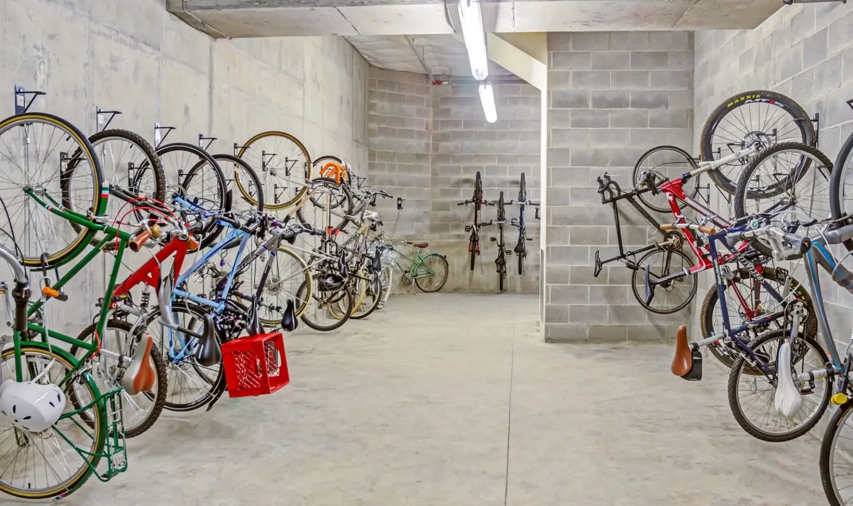 Concrete room with bicycles mounted on walls, well-lit by fluorescent lights. Various bikes, including road and mountain bikes, are securely stored.