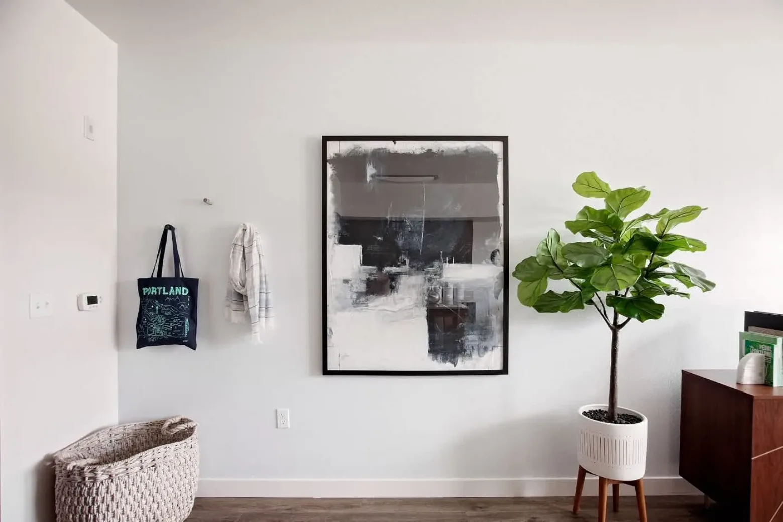 A minimalist apartment interior featuring a large abstract painting on the wall, a hanging tote bag, a plaid towel, a woven basket, and a potted fiddle leaf fig tree on a wooden stand.
