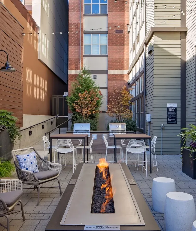 A cozy outdoor courtyard with a central fire pit, seating, and decorative plants, nestled between modern buildings and adorned with string lights.