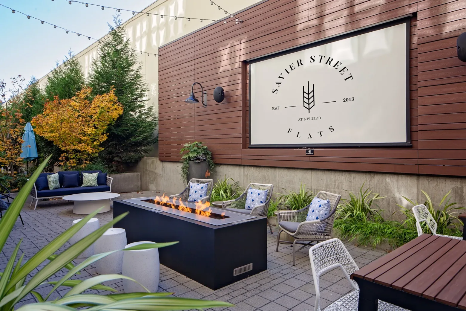 Modern outdoor space with fire pit, seating, and greenery at Savier Street Flats. String lights create an inviting atmosphere. No people present.