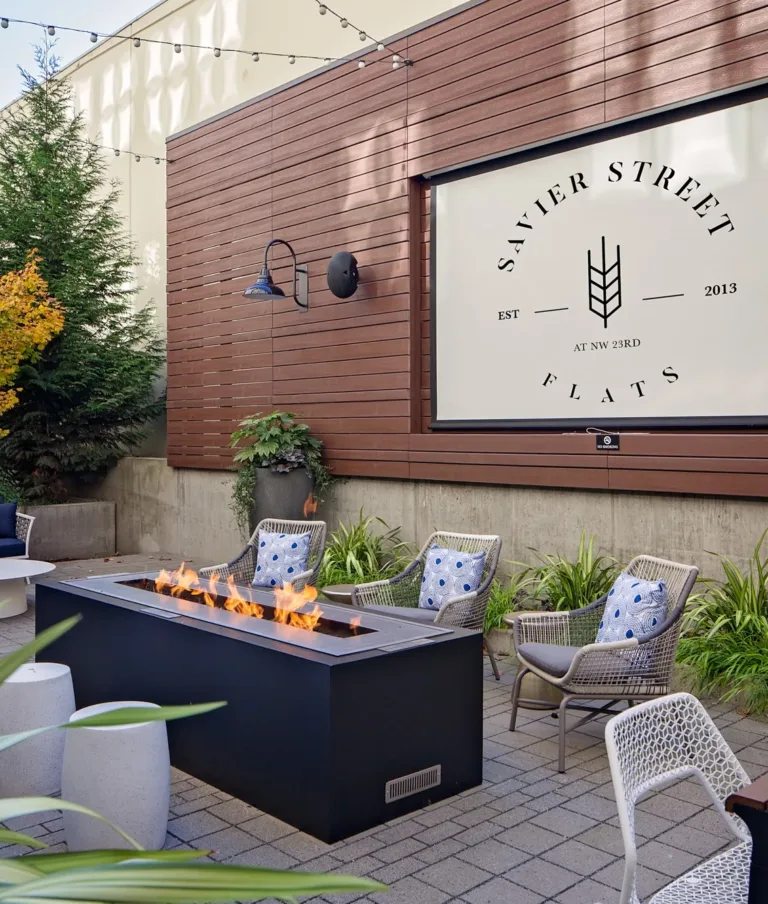 Modern outdoor space with fire pit, seating, and greenery at Savier Street Flats. String lights create an inviting atmosphere. No people present.