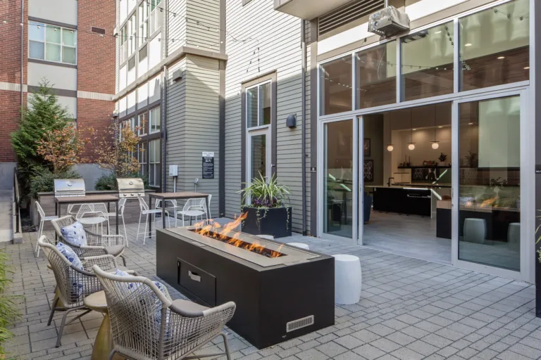 Modern outdoor patio with fire pit, seating, and grills. Adjacent to building entrance featuring large windows, contemporary design, and neutral colors. Cozy ambiance.