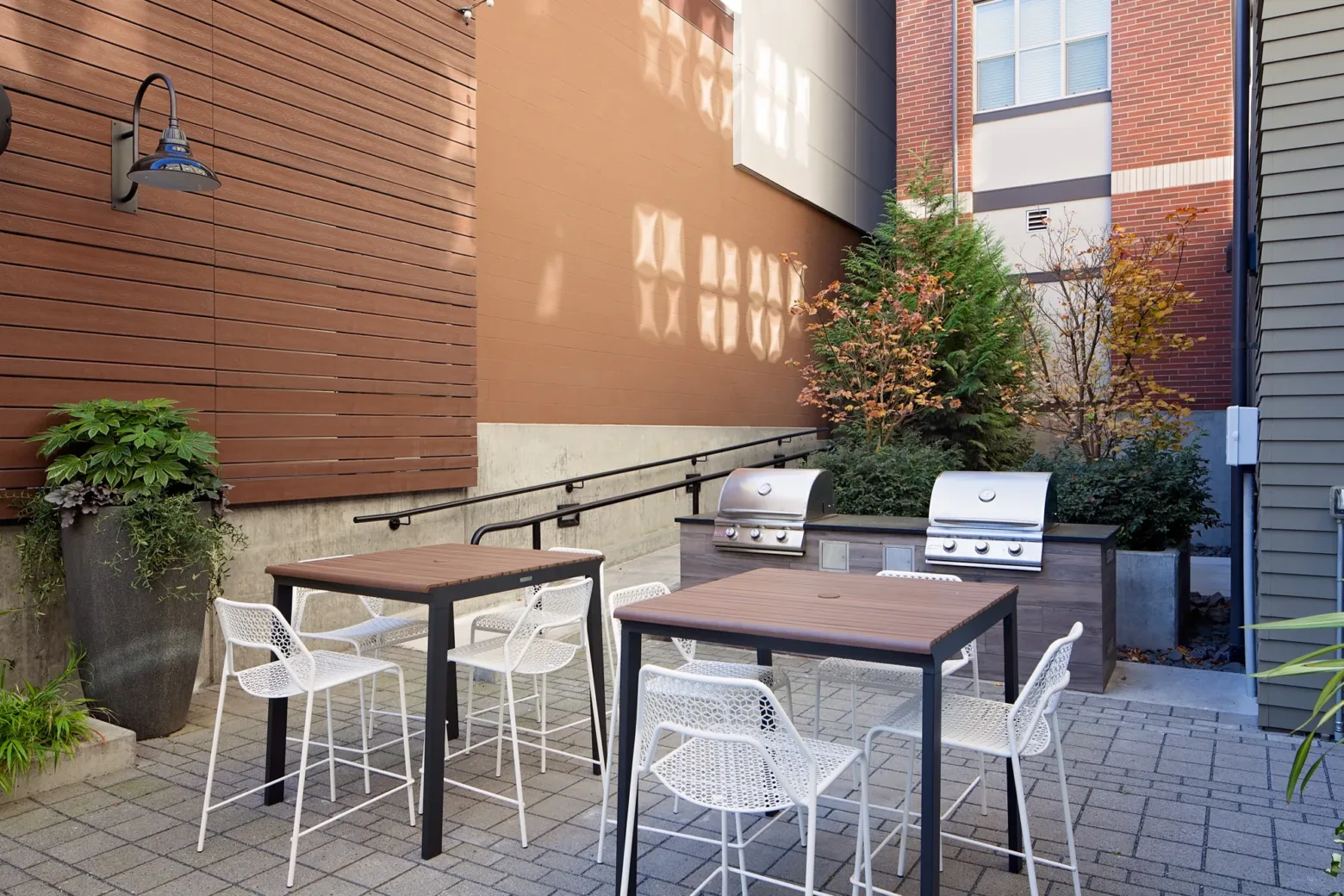 Outdoor patio with two grill stations, wooden tables, white chairs, and potted plants, surrounded by modern brick and wooden building facades.