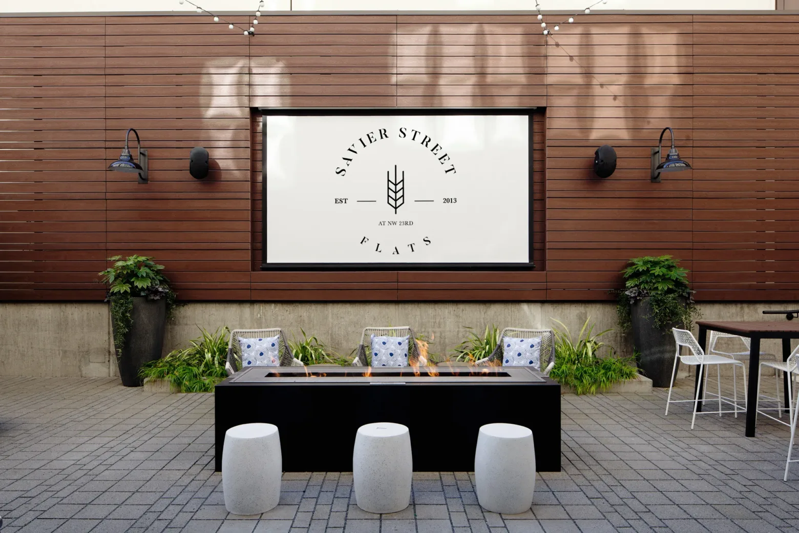 Modern outdoor lounge area with a fire pit, stools, and plants. "Savier Street Flats" logo on wood-paneled wall, featuring string lights overhead.