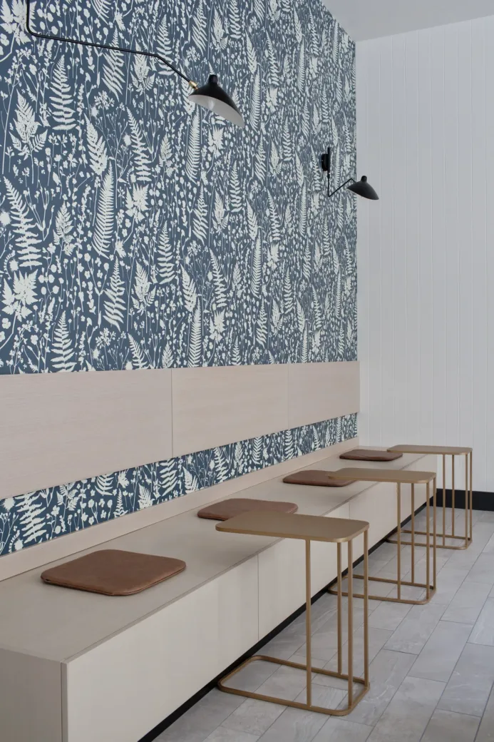A modern community room with a wall featuring dark blue and white floral wallpaper, minimalist wooden benches with brown cushions, and sleek metal side tables beneath black adjustable wall lamps.