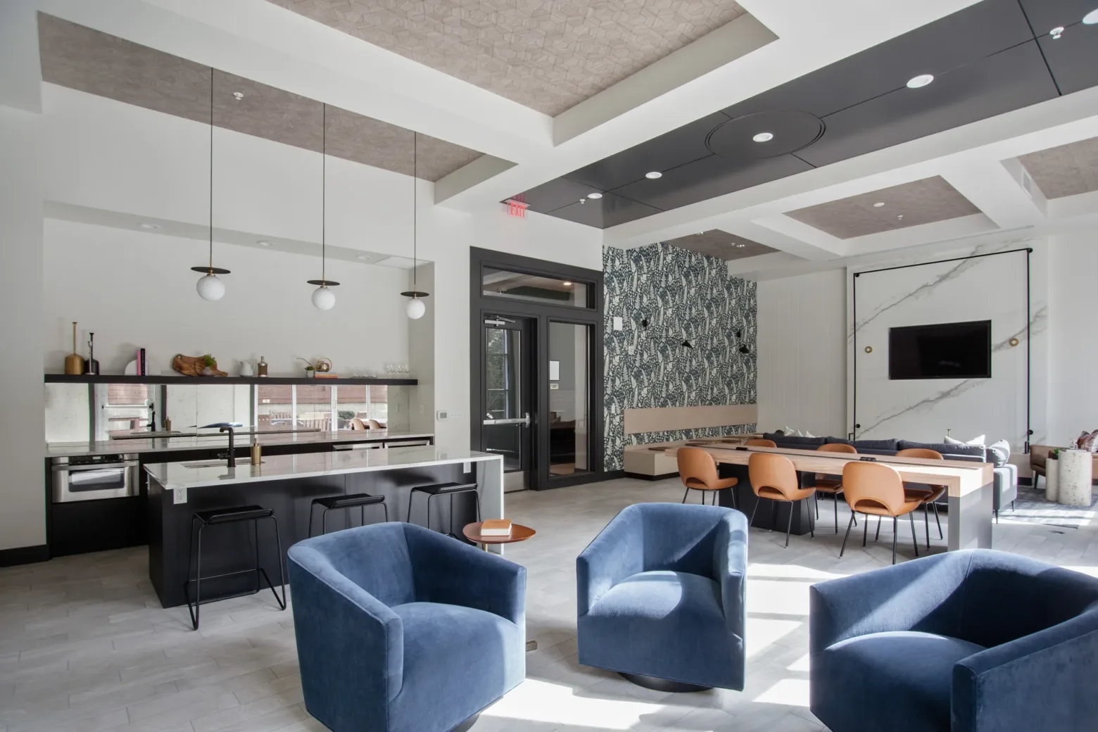 A modern community room featuring blue velvet swivel chairs, a sleek kitchen with white countertops and pendant lights, a large wooden table with tan chairs, and a wall-mounted TV. The room has patterned wallpaper and geometric ceiling details, with abundant natural light from windows.