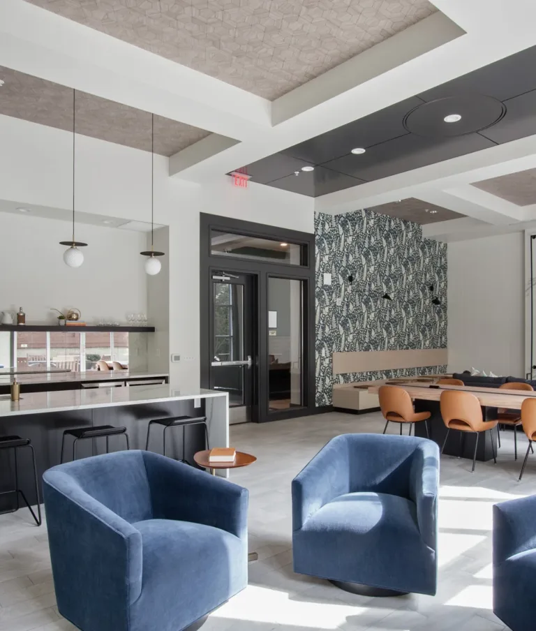 A modern community room featuring blue velvet swivel chairs, a sleek kitchen with white countertops and pendant lights, a large wooden table with tan chairs, and a wall-mounted TV. The room has patterned wallpaper and geometric ceiling details, with abundant natural light from windows.