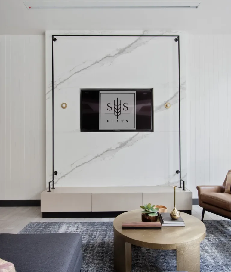A modern community room with a wall-mounted TV between white marble panels, a comfortable brown leather armchair with a patterned cushion, a round wooden coffee table with succulents and books, and a dark blue sofa. The room features a blue and white patterned wall and wooden flooring.