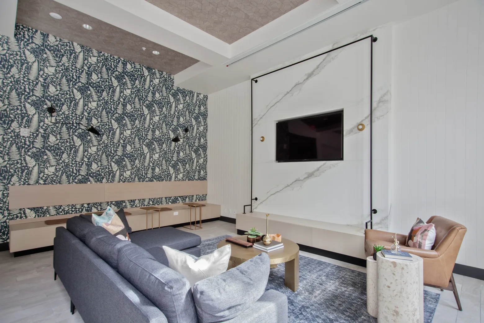 A modern community room featuring a dark gray sofa, a brown leather armchair, and a round wooden coffee table on a textured rug. The room has a wall-mounted flat-screen TV on a marble accent wall, a patterned wallpaper with abstract designs, and overhead lighting fixtures. Decorative items are placed on the table and side tables.