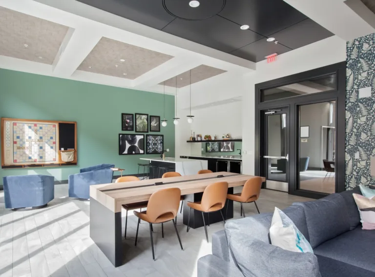 A modern community room featuring a green accent wall with art, a large wooden table with brown chairs, a blue sofa, and armchairs. The room includes a kitchenette with black cabinets, a light fixture with hanging spheres, and a large Scrabble board on the wall.