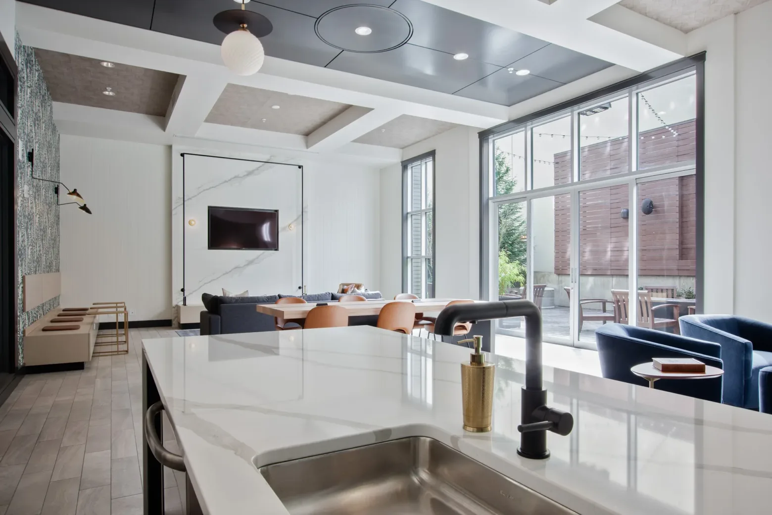 A modern community room featuring a marble-topped island with a black faucet, a sitting area with a large TV mounted on a marble accent wall, leather chairs, a couch, and large windows allowing natural light to fill the space.