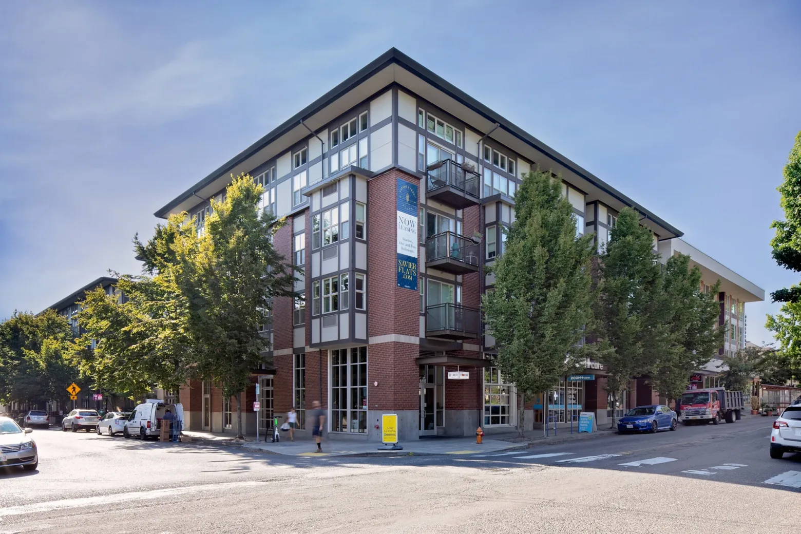 A multi-story brick and glass building with balconies, surrounded by trees and positioned on a street corner with cars parked along the street.