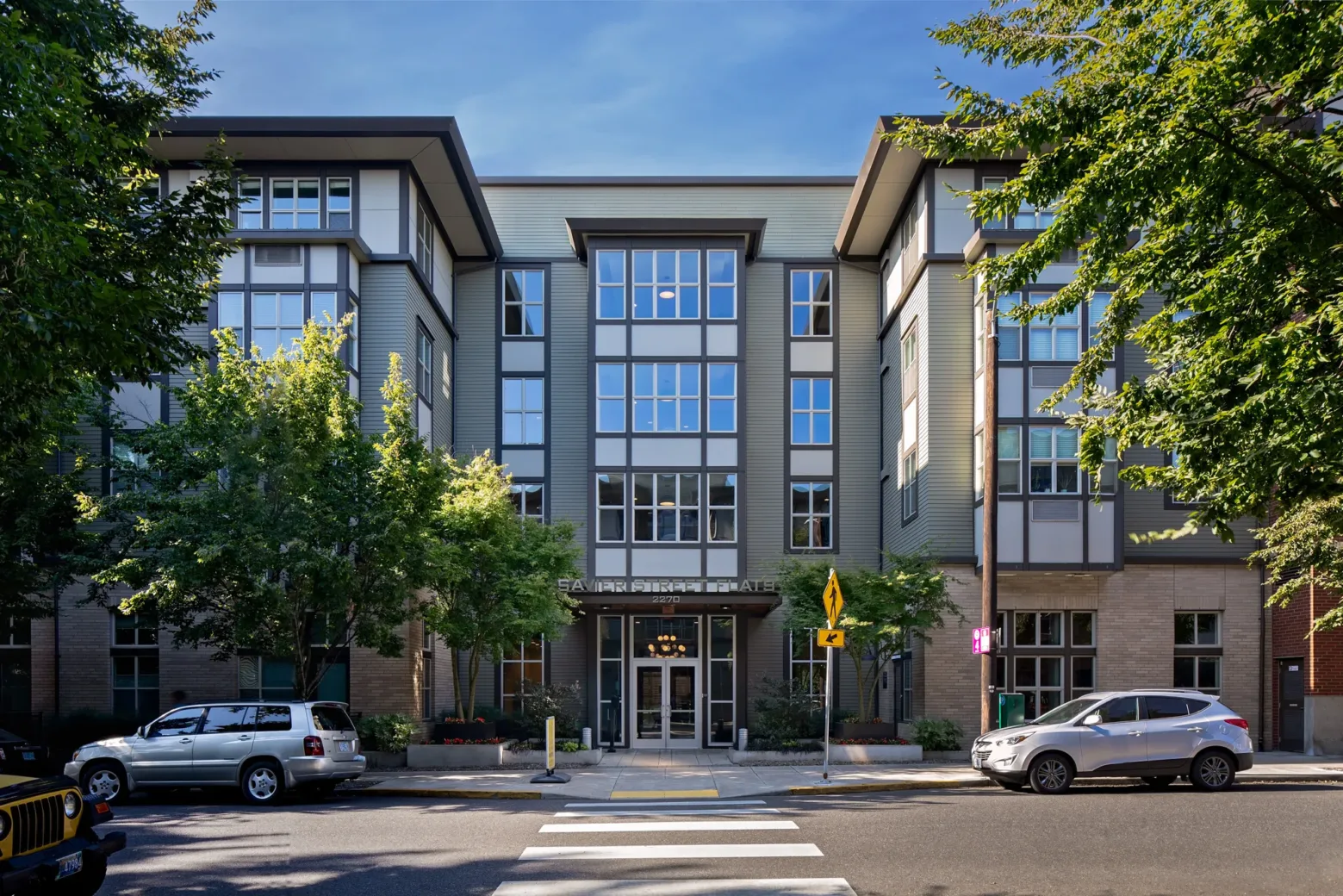 A four-story residential building with modern architecture, featuring large windows and surrounded by trees, with parked cars on the street in front.
