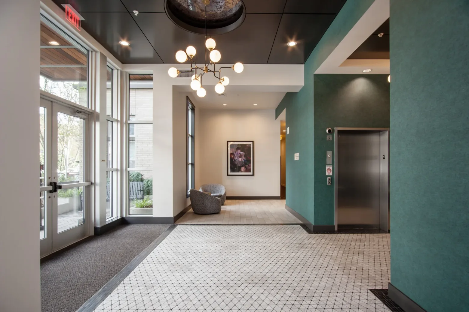 A modern lobby with turquoise walls, elevator, decorative chandelier, large windows, and a framed artwork on the wall above a cozy chair.