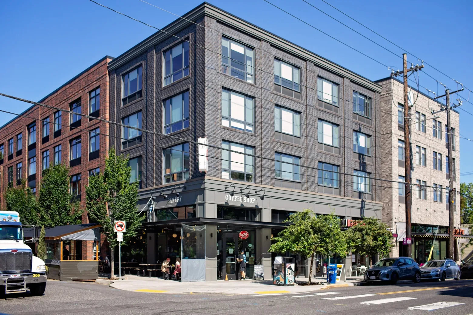 A modern four-story building with a coffee shop on the ground floor, people sitting outside, and parked cars on the city street.