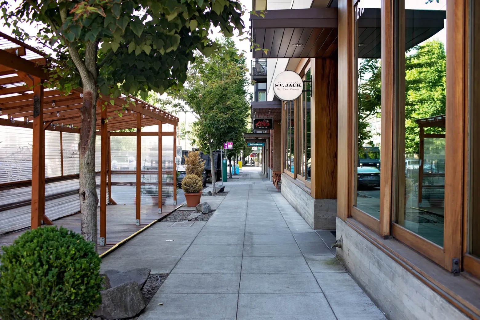 A tree-lined sidewalk with storefronts, including "St. Jack" restaurant, outdoor seating area, and parked cars. Peaceful urban setting with lush greenery.