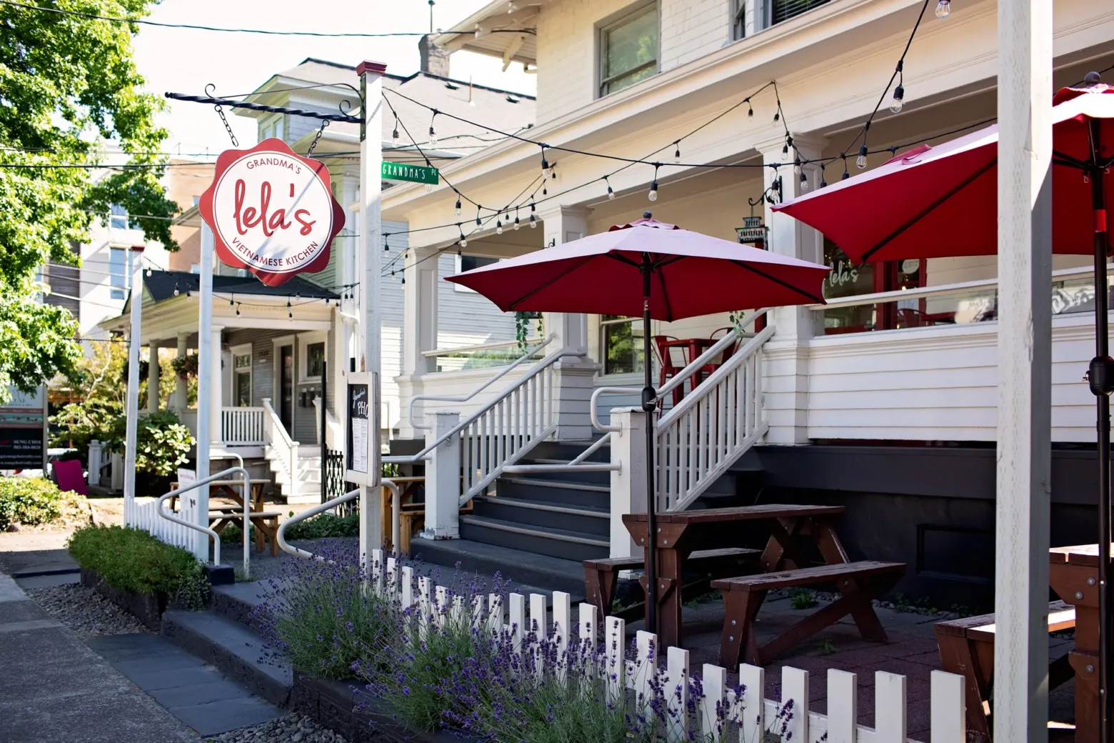 A cozy outdoor dining area with red umbrellas at Lela’s Vietnamese Kitchen, featuring white picket fence and string lights in an urban setting.