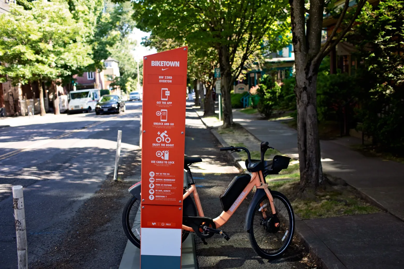 A Biketown rental station with an orange bike is on a tree-lined street, featuring parked cars and residential homes in the background.