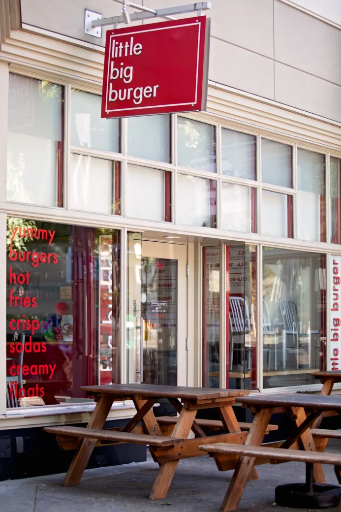 Small burger restaurant with a red sign, featuring outdoor wooden picnic tables. Glass windows display menu items like burgers and sodas. Quiet street scene.