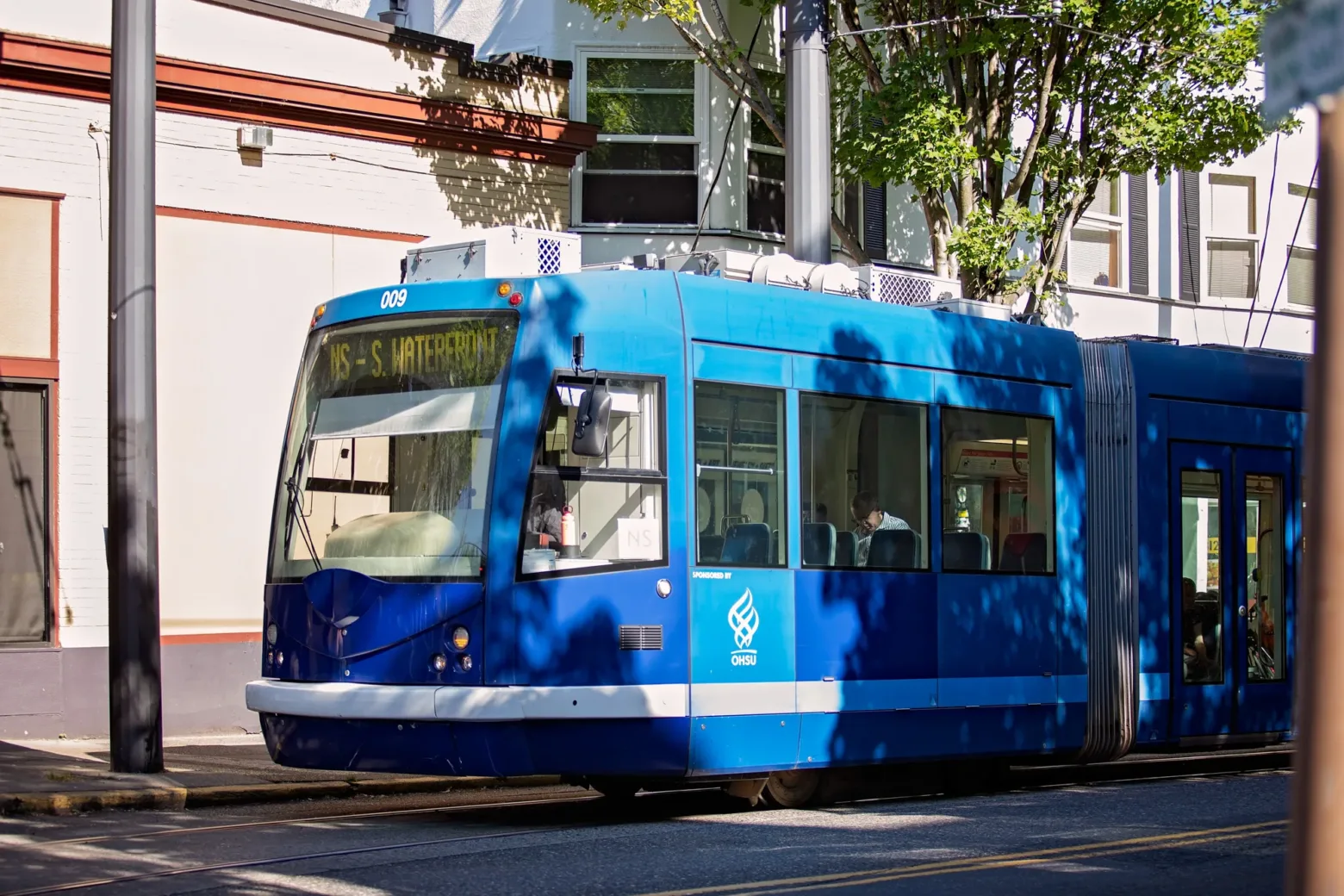 A blue tram labeled "S. Waterfront" travels through a tree-lined street with people inside. Sunlight casts shadows on nearby buildings.