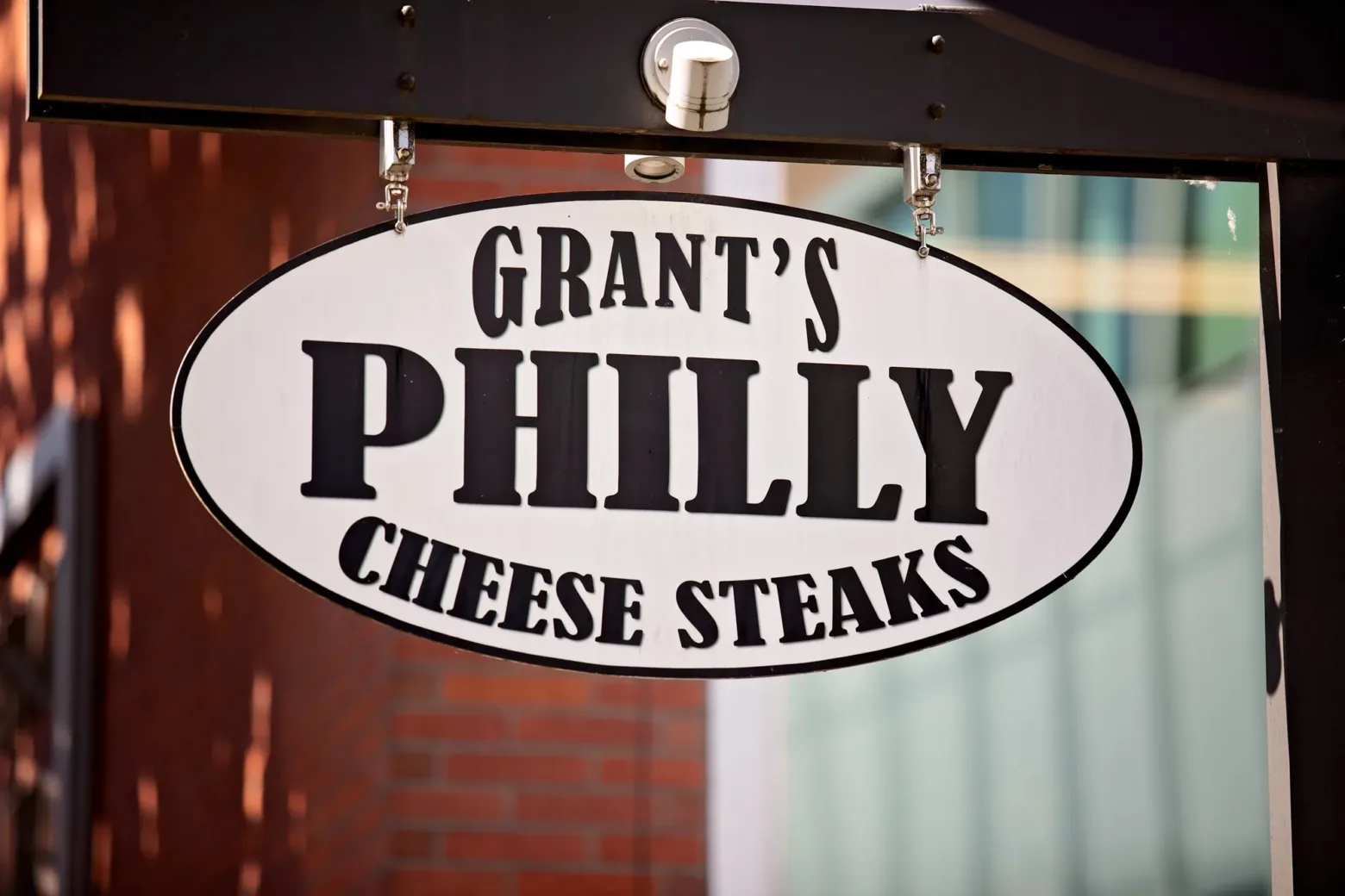 Oval sign for "Grant's Philly Cheese Steaks" hangs from a bracket, with blurred red brick wall in the background.