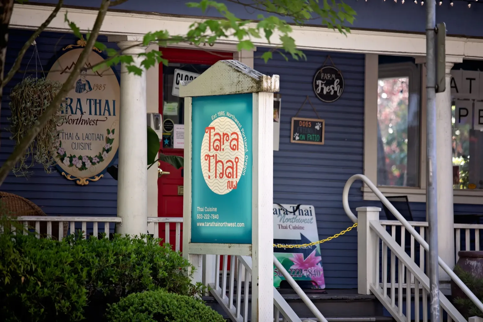 A Thai restaurant named "Tara Thai Northwest" in a blue house with a porch, surrounded by greenery. Signage indicates Thai and Laotian cuisine.