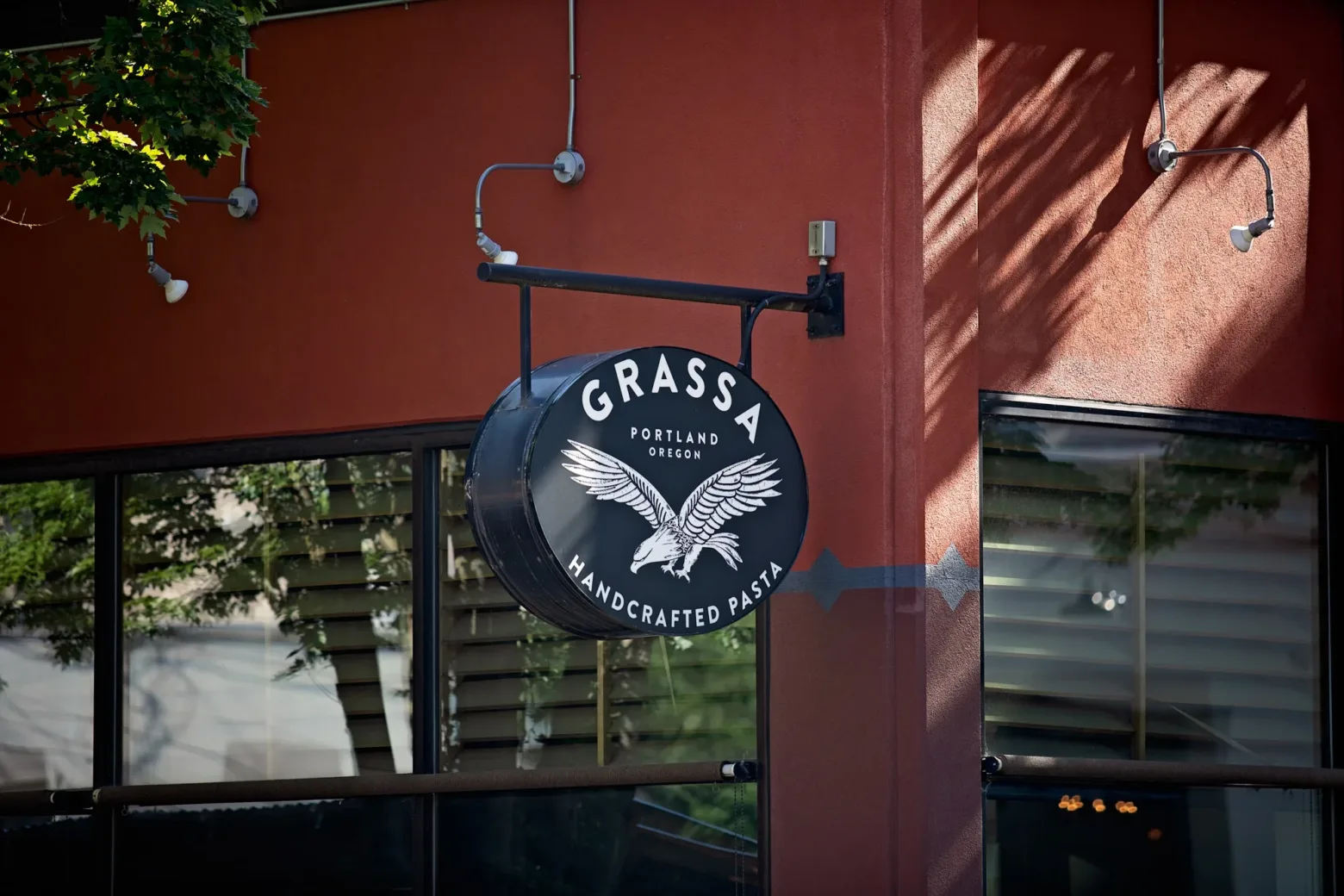 Outdoor sign for "Grassa Handcrafted Pasta" in Portland, Oregon, displayed on a brick wall with a tree shadow above.