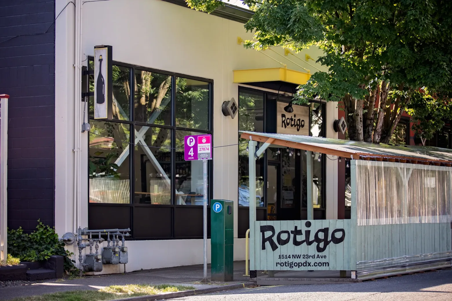 Restaurant "Rotigo" with glass windows and outdoor seating under trees, on a corner street with parking signs and a wine bottle logo.