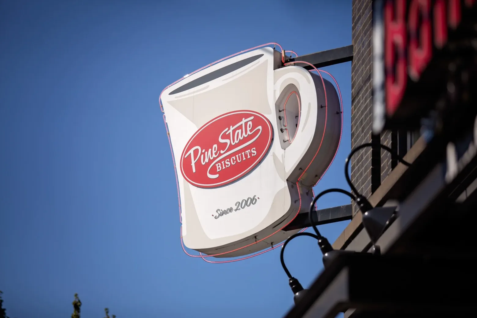 A large coffee cup sign for "Pine State Biscuits" hangs on a building exterior against a clear blue sky, established in 2006.