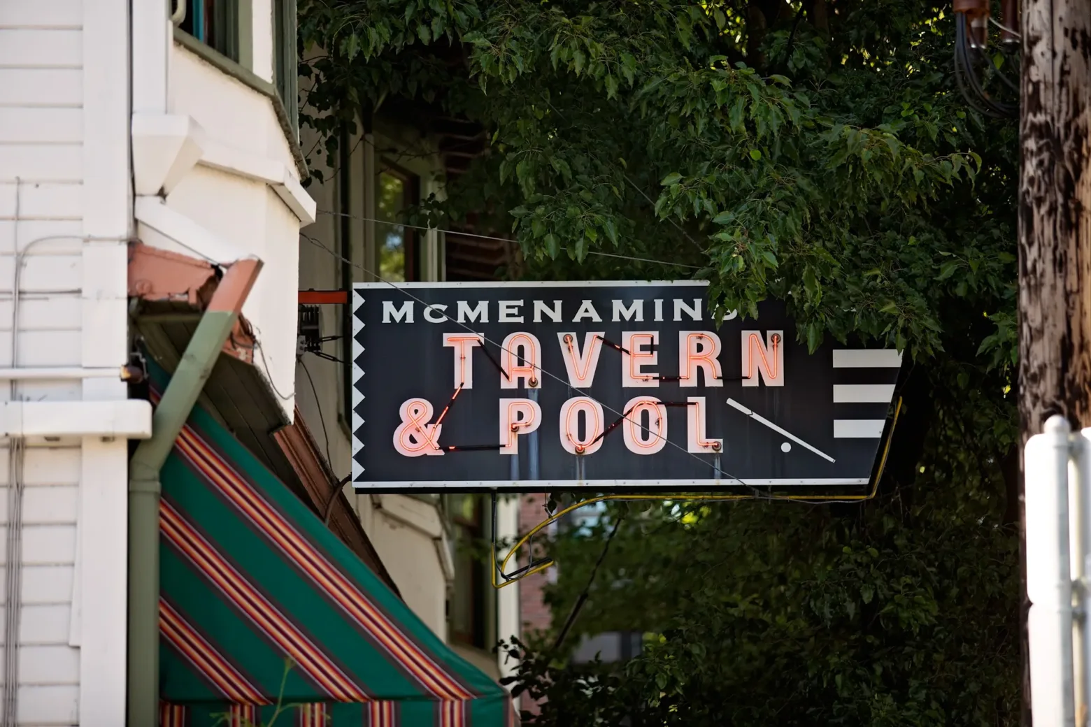 A McMenamin's Tavern & Pool sign hangs near a building with a striped awning, surrounded by green foliage on a sunny day.