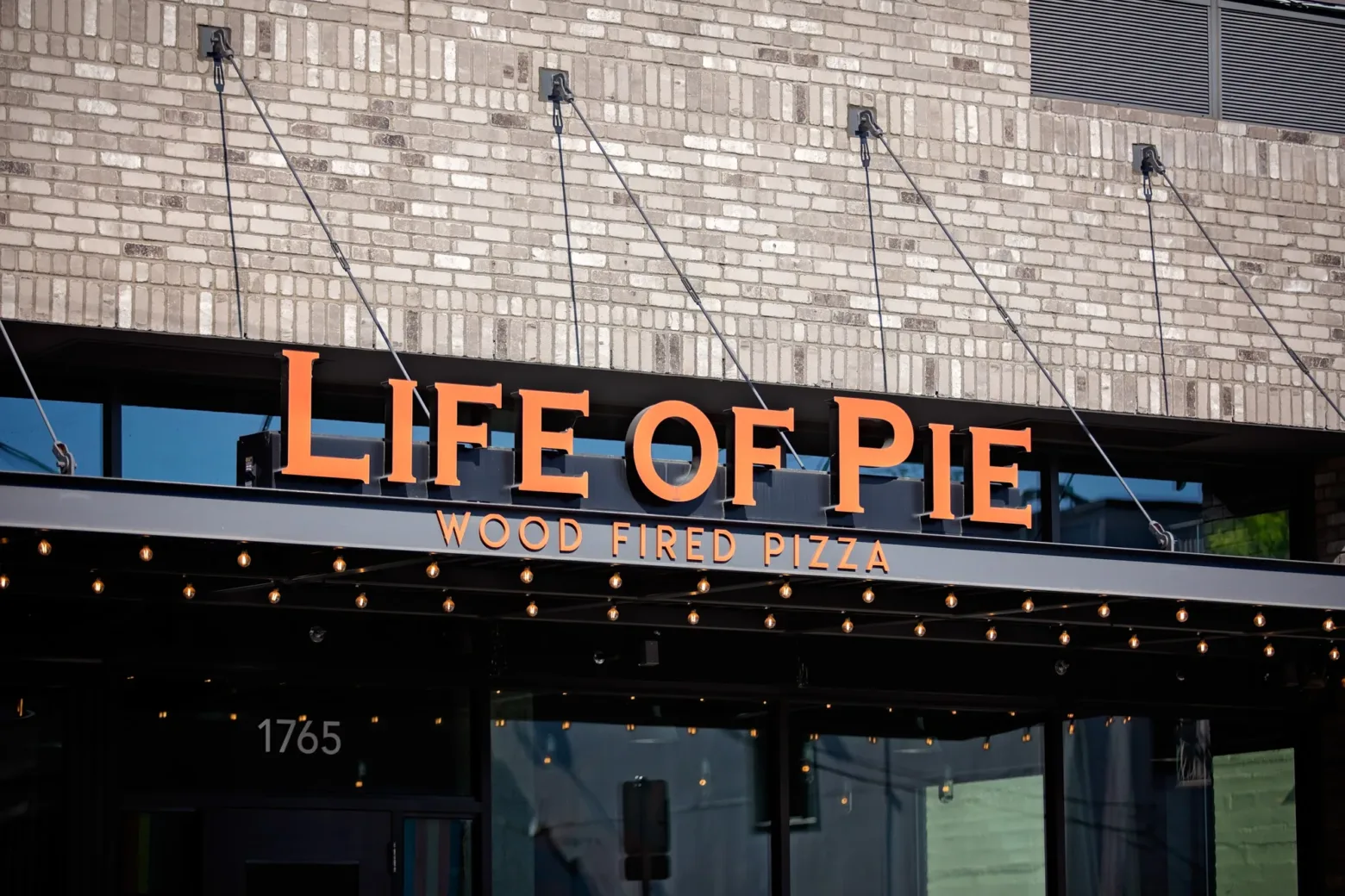 Restaurant storefront with "Life of Pie" sign, offering wood-fired pizza. Brick exterior, decorative lights, address number 1765 displayed prominently.
