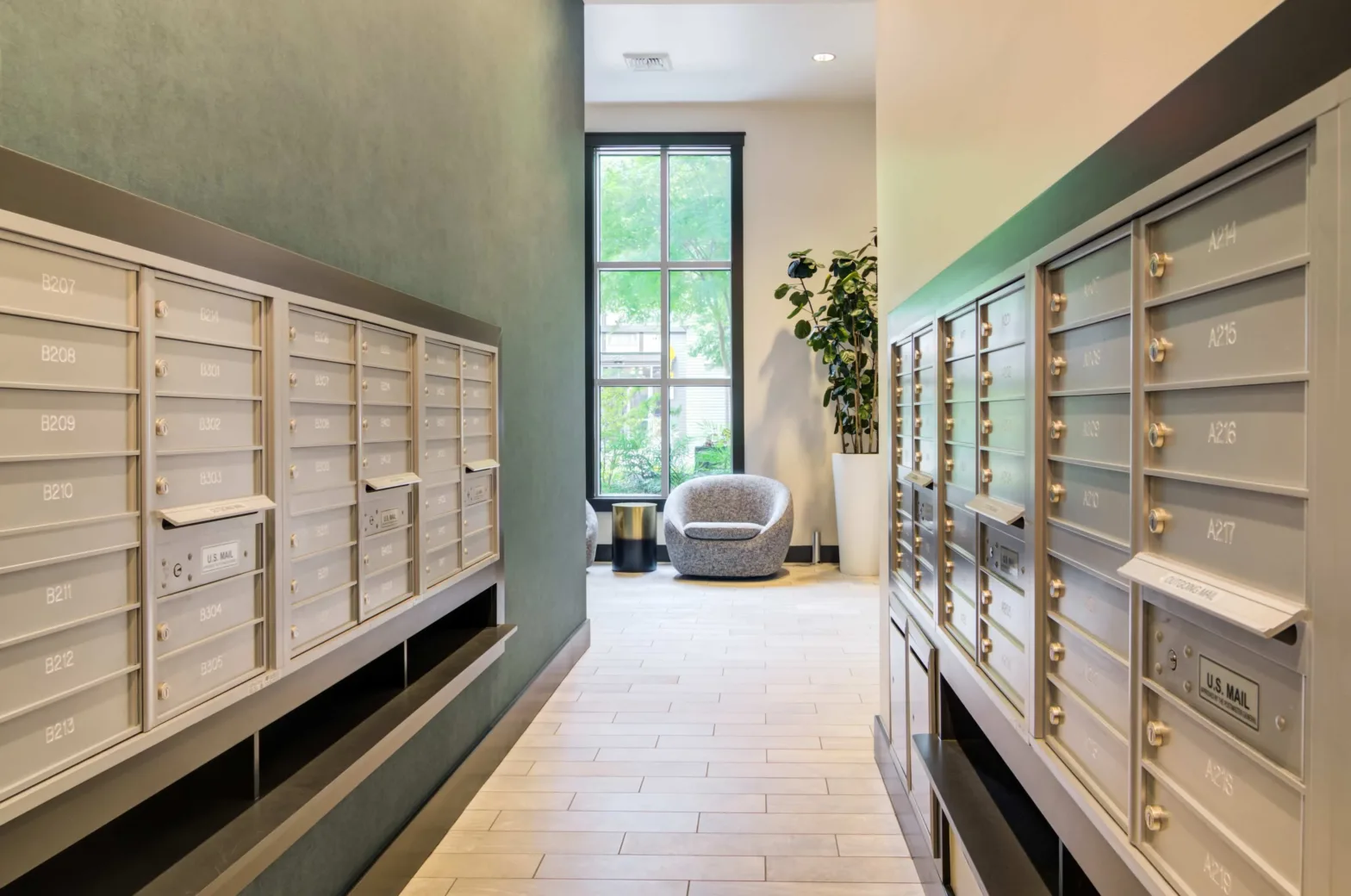 A hallway features gray mailboxes lining both walls, a window with natural light, a gray chair, and a tall potted plant.