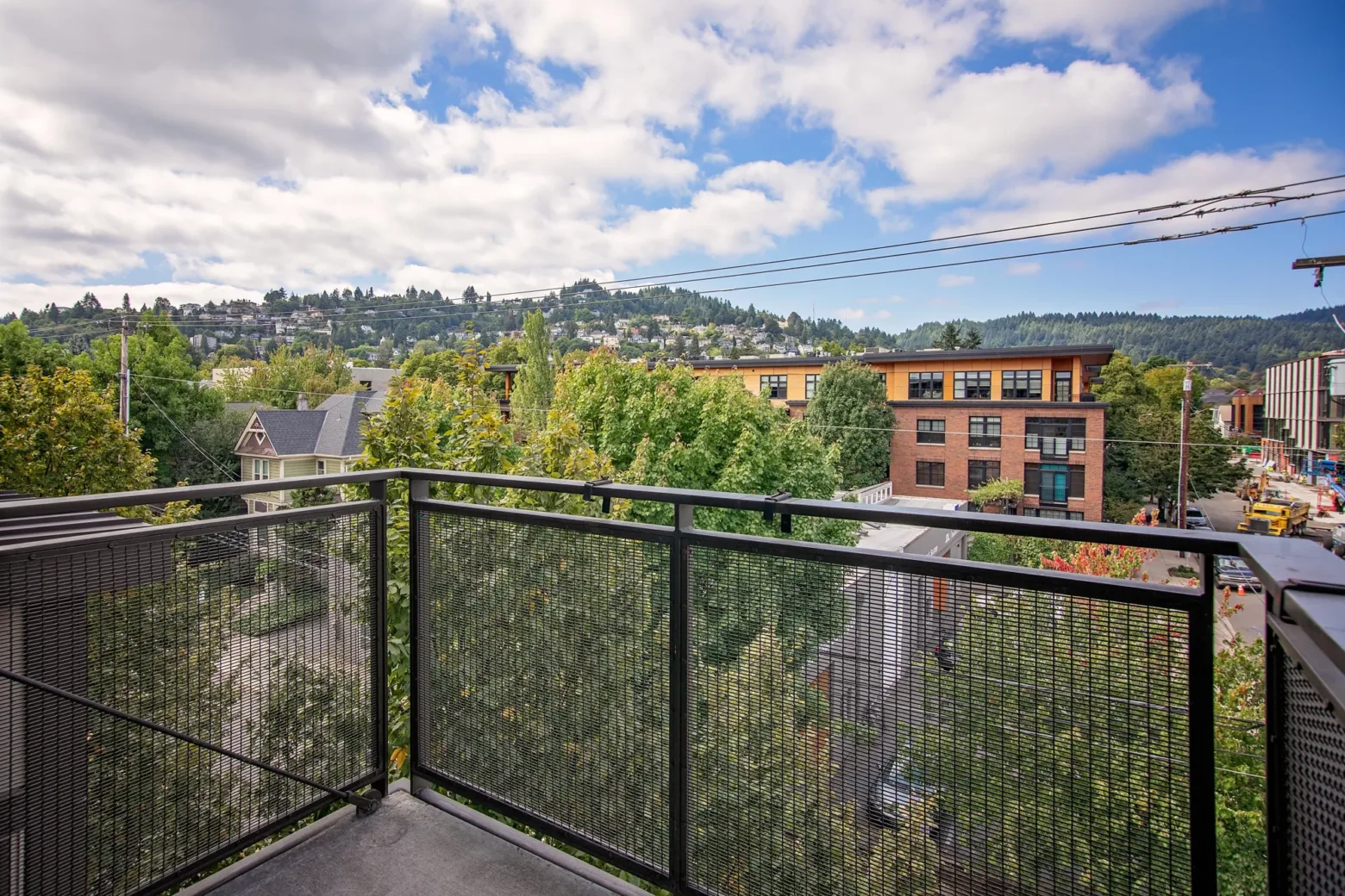 A view from a balcony with a mesh railing overlooking lush green trees and a mix of residential and brick buildings under a partly cloudy blue sky.