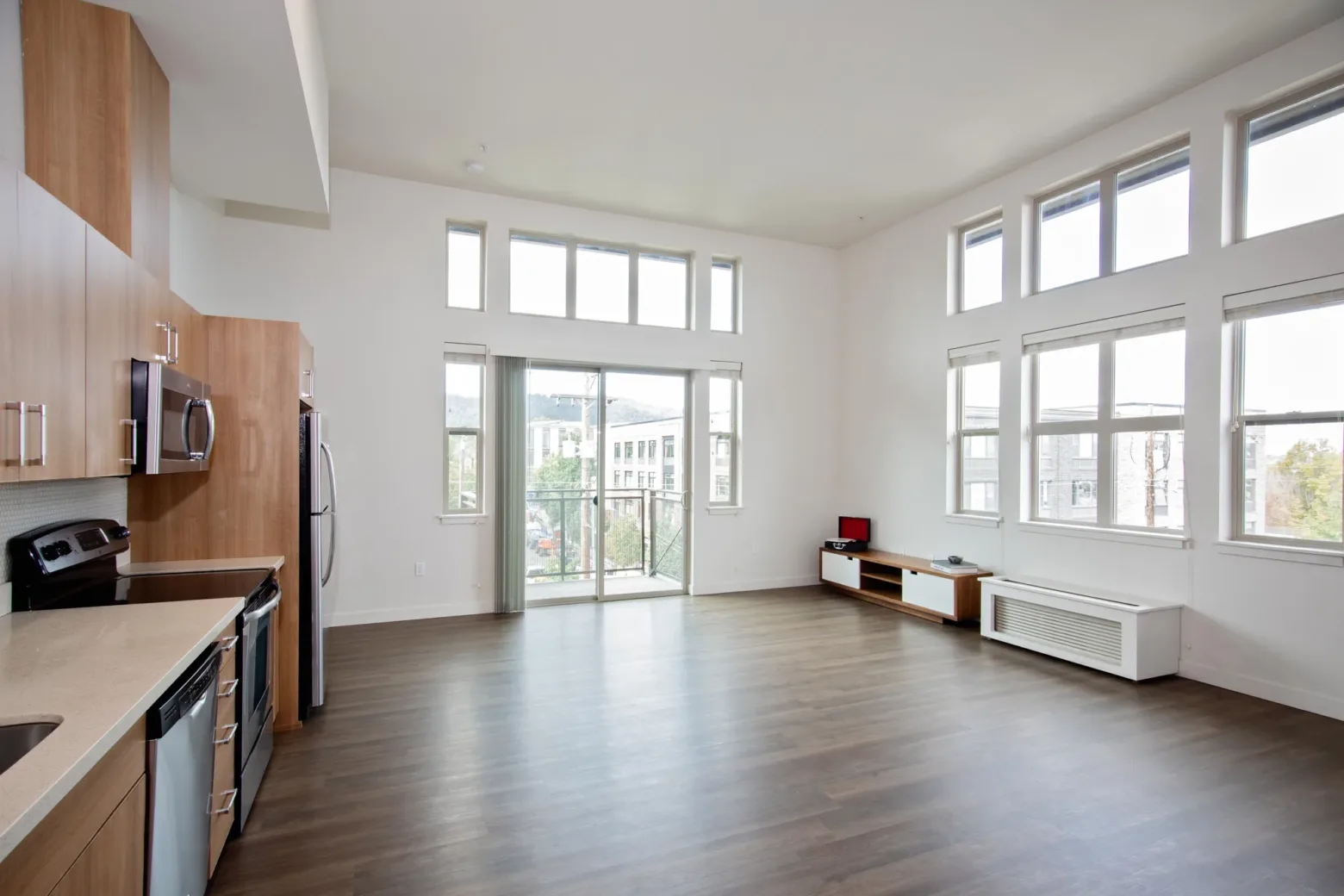 Spacious apartment interior with large windows, light wood flooring, and an open kitchen that includes stainless steel appliances and wooden cabinets.