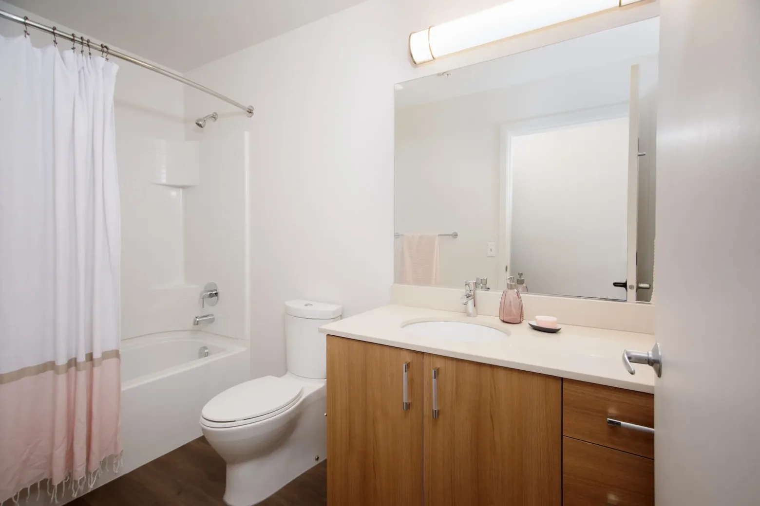 Modern apartment bathroom with a white shower curtain, wooden vanity, and white fixtures.