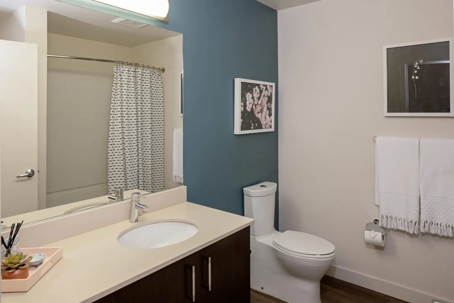 A modern bathroom with a dark wood vanity and white countertop, featuring a sink and faucet. A toilet is adjacent to a blue accent wall with floral artwork. There is a shower with a patterned curtain and white towels hanging on the wall.