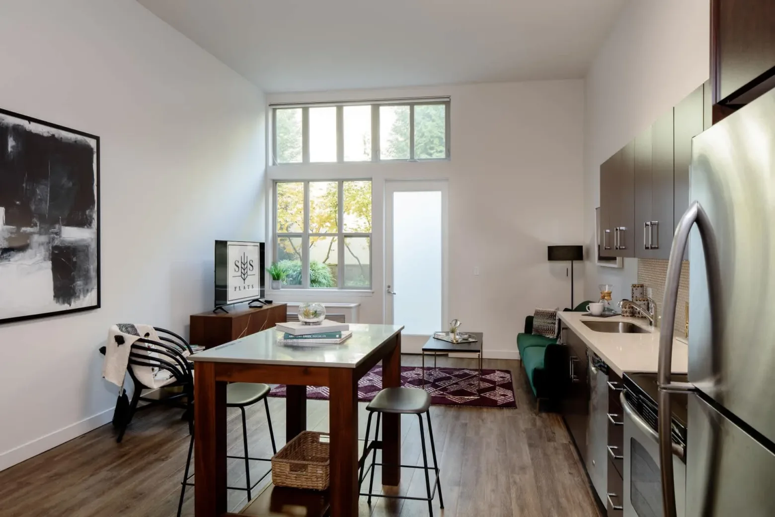A modern, open-plan apartment living area with a kitchen and sitting space. The room features wooden flooring, a high ceiling with large windows, and a minimalist aesthetic. Furniture includes a wooden dining table with stools, a small green sofa, and a black chair. Appliances are stainless steel, and there's contemporary art on the wall.