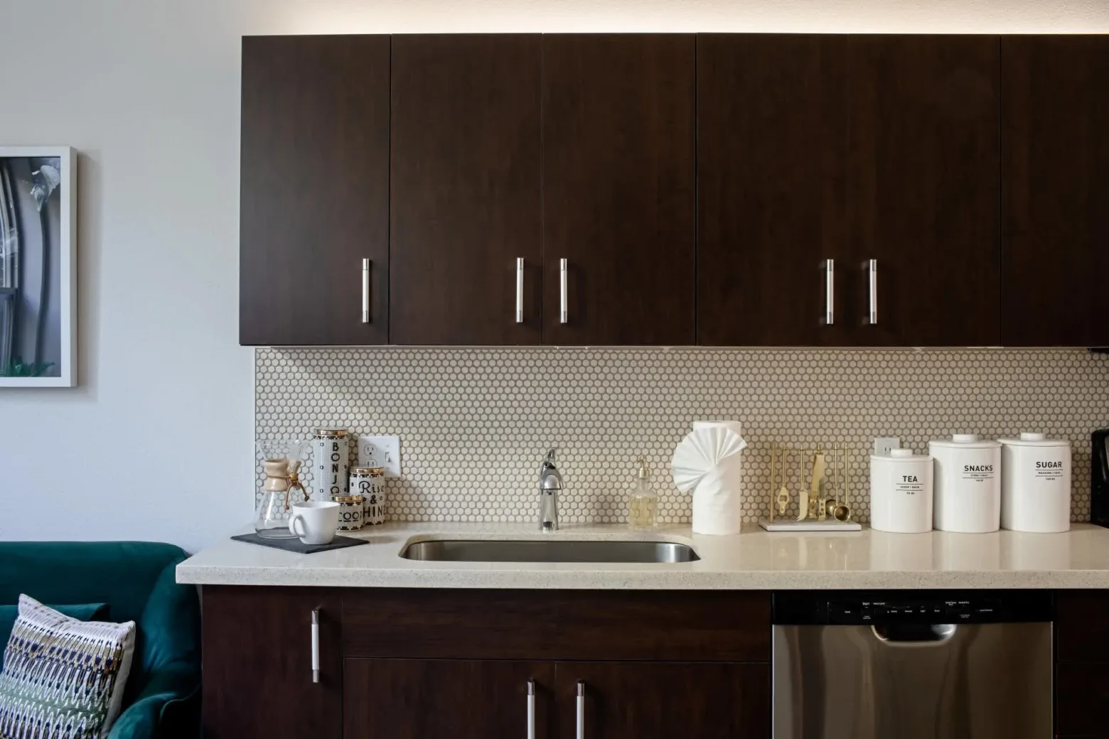 A modern kitchen with dark wooden cabinets, a light countertop, and a stainless steel sink. There are white containers labeled 'Tea,' 'Snacks,' and 'Sugar' on the counter, along with a roll of paper towels and jars of dry goods. A backsplash with small, circular tiles complements the sleek design.