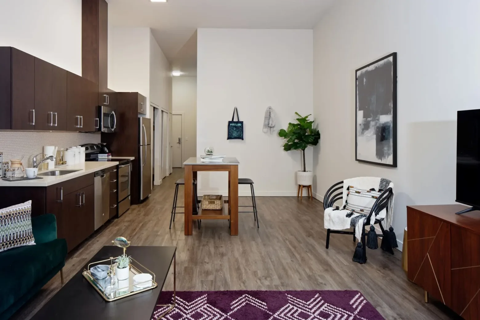 Modern apartment interior with an open-plan layout featuring a kitchen on the left with dark cabinets, stainless steel appliances, and a dining area with a tall wooden table and stools. The living area includes a green sofa, coffee table with decor, and a chair in front of a large wall art piece. A potted plant and wall-mounted decor add a touch of style, with a wood floor tying the space together.
