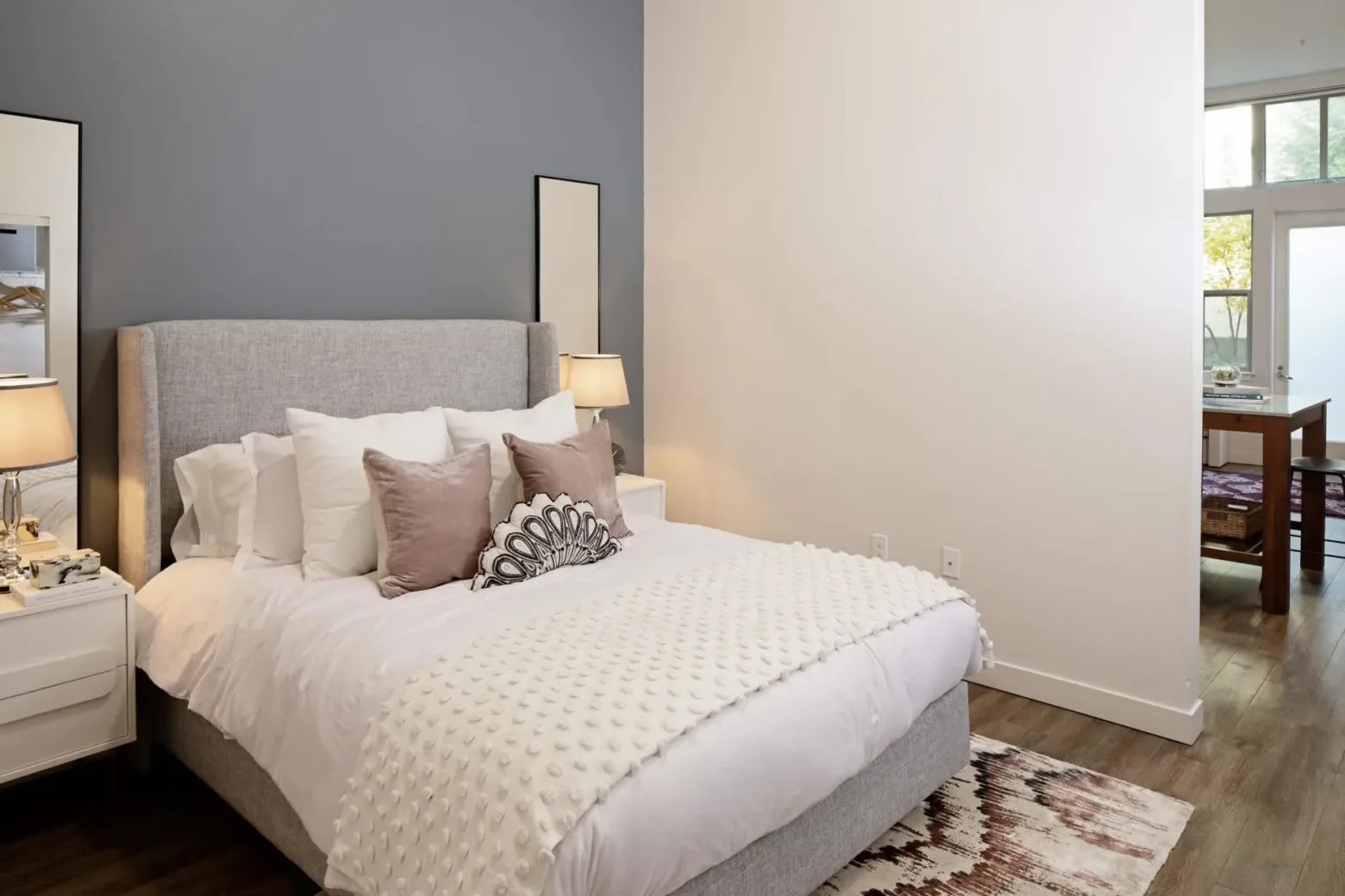 Modern bedroom with a gray upholstered bed featuring white and beige pillows, a textured white blanket, and matching side tables with lamps against a two-tone wall.