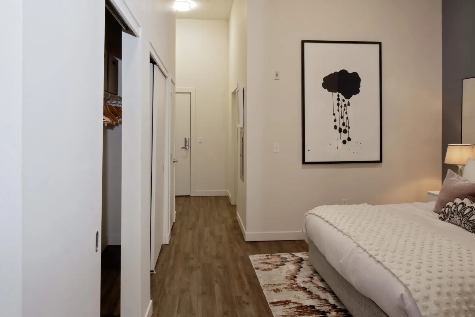 A modern apartment bedroom featuring a bed with a textured white comforter, a brown and white patterned rug, and a bedside lamp. A large abstract art piece hangs on the wall, and there are closets with sliding doors along the hallway leading to the entrance.