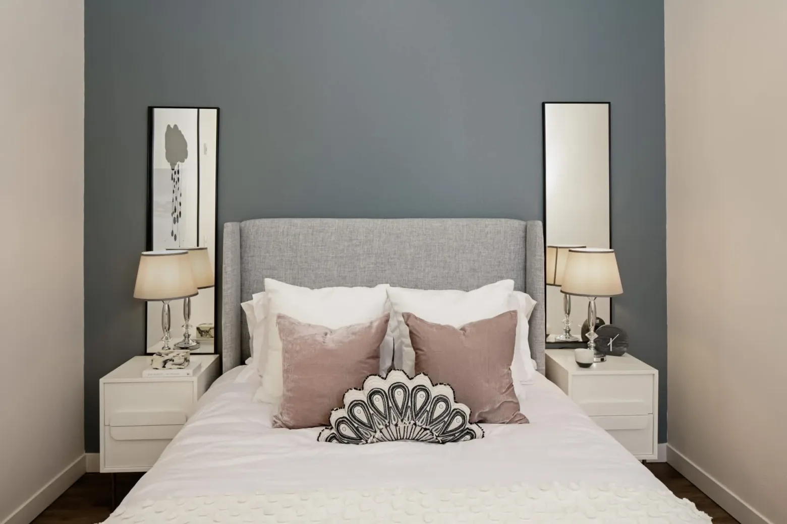 Modern bedroom with a gray upholstered bed, decorative pillows, white nightstands with lamps, and a blue-gray accent wall.